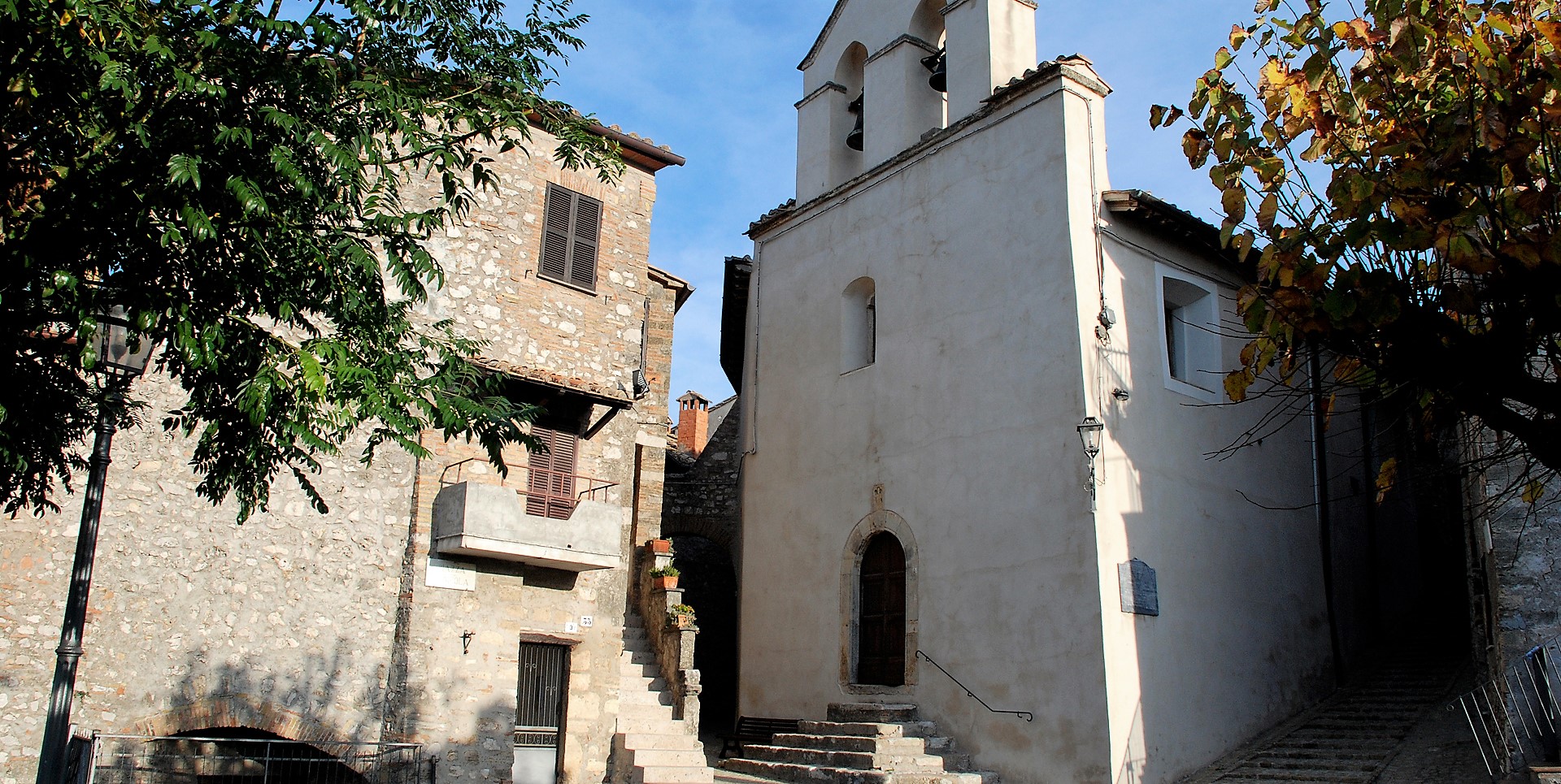 Church with a bell-gable and stone staircase, next to a stone house with shutters and a flowered balcony.