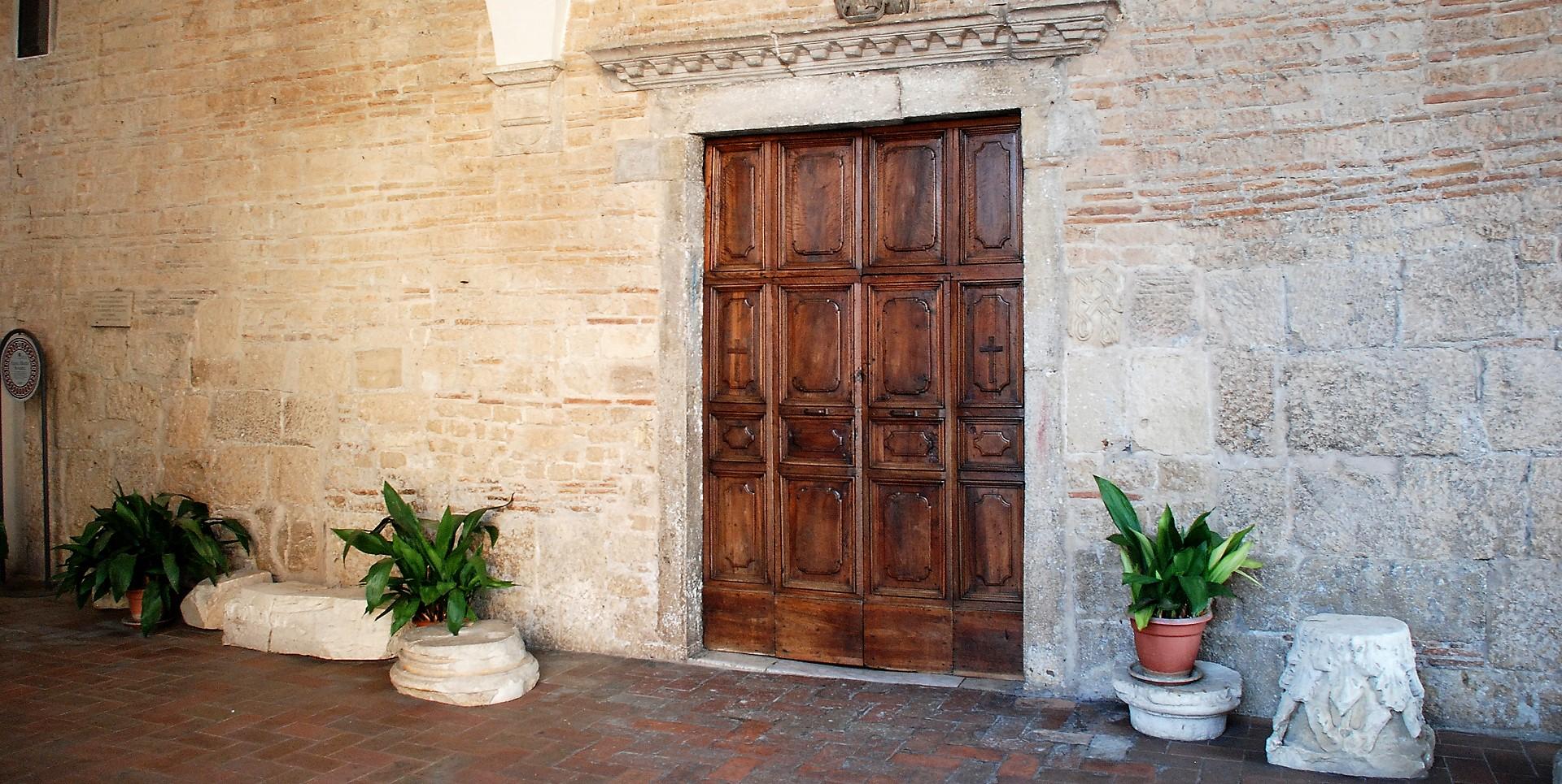 Portale ligneo della Collegiata, incorniciato da mura in pietra e affiancato da piante in vaso.