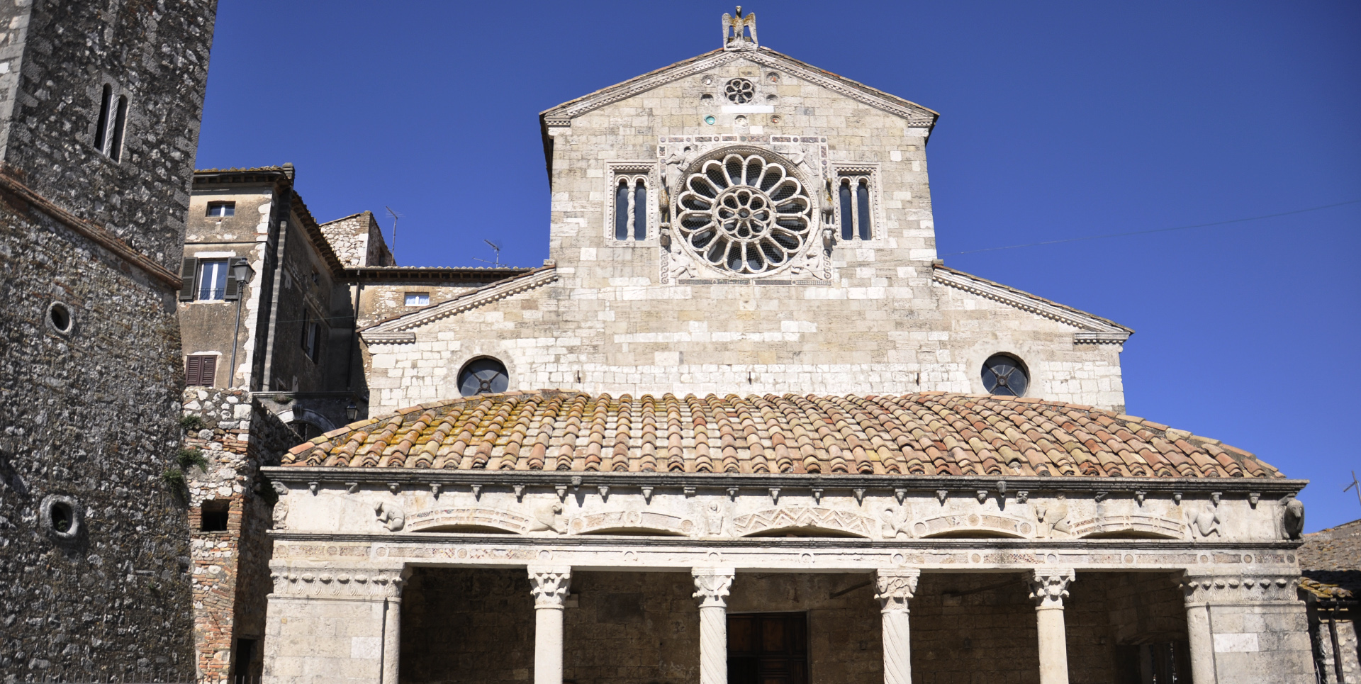 Facciata in pietra della chiesa di Santa Maria Assunta con rosone centrale e portico a colonne sotto cielo azzurro.