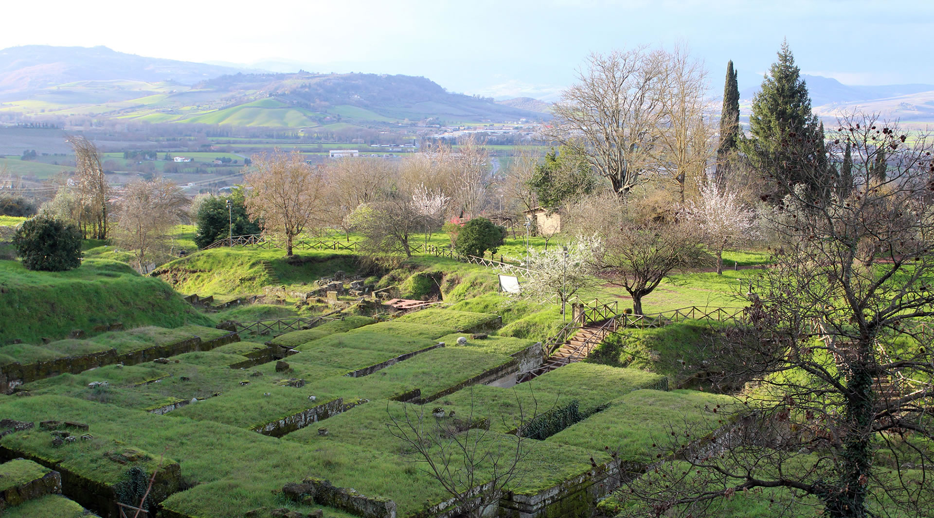 Aerial view of the Etruscan necropolis with tumulus tombs topped with grassy roofs, surrounded by hills.