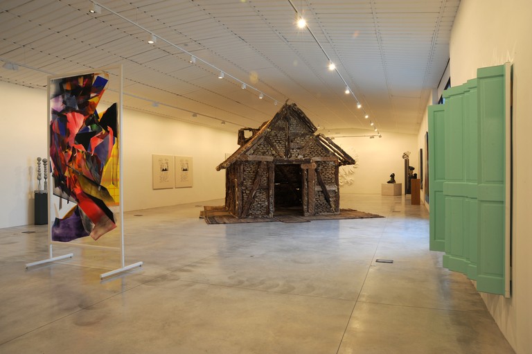 An exhibition room with a sculpture of a hut in the center, an abstract artwork on the left, and a green window on the right.