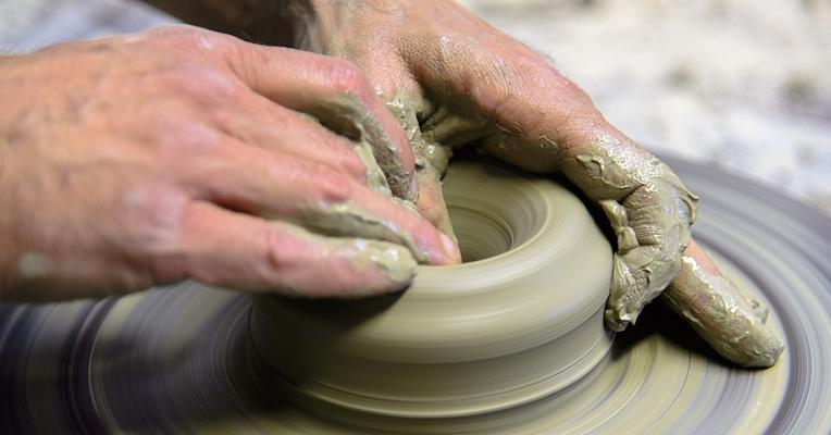 Hands shaping clay on a spinning pottery wheel, forming the neck and rim of a vase