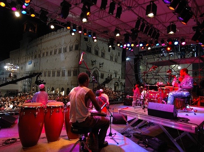 Vista dal palco di un concerto di Umbria Jazz a Perugia, con musicisti che suonano strumenti a percussione davanti al pubblico, illuminati da luci colorate, con il Palazzo dei Priori sullo sfondo.