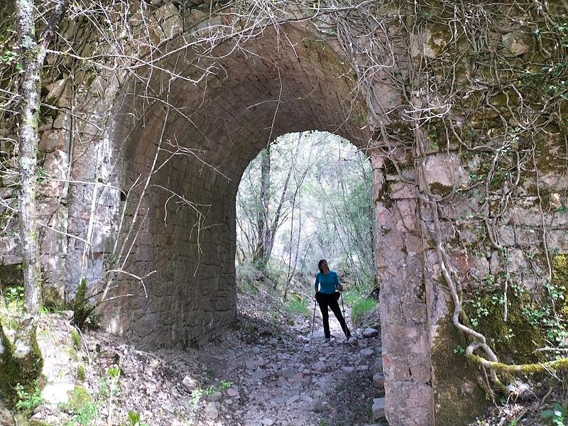 Escursionista sotto un antico ponte in pietra, con muschi e piante rampicanti, immerso  nei boschi dei Monti Martani, nei pressi di Macerino
